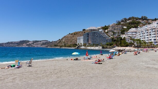 A beach with a clear blue sky, sandy shore, and buildings in the background.