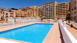 A swimming pool with a wooden deck and white railings, surrounded by multi-story buildings with balconies.