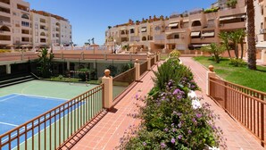 A courtyard with a tennis court, surrounded by multi-story buildings with balconies and a garden with purple flowers.