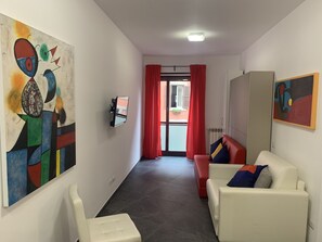 Un couloir avec un canapé blanc, un canapé rouge et un fauteuil blanc. Les murs sont ornés de tableaux abstraits et un téléviseur est fixé au mur.