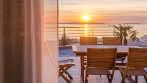 A terrace with a wooden table and chairs, overlooking a sunset over the water.