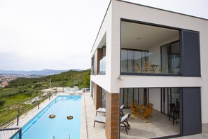 A modern two-story building with a swimming pool, outdoor seating, and a view of the countryside. - Royal Villa 2 (Podstrana)