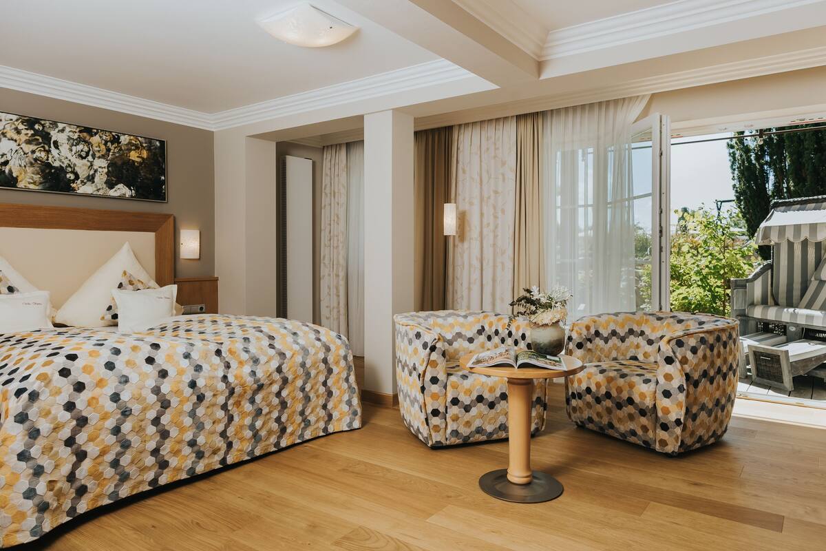 A hotel room with a large bed, two patterned armchairs, a small round table, and a view of the outdoors.