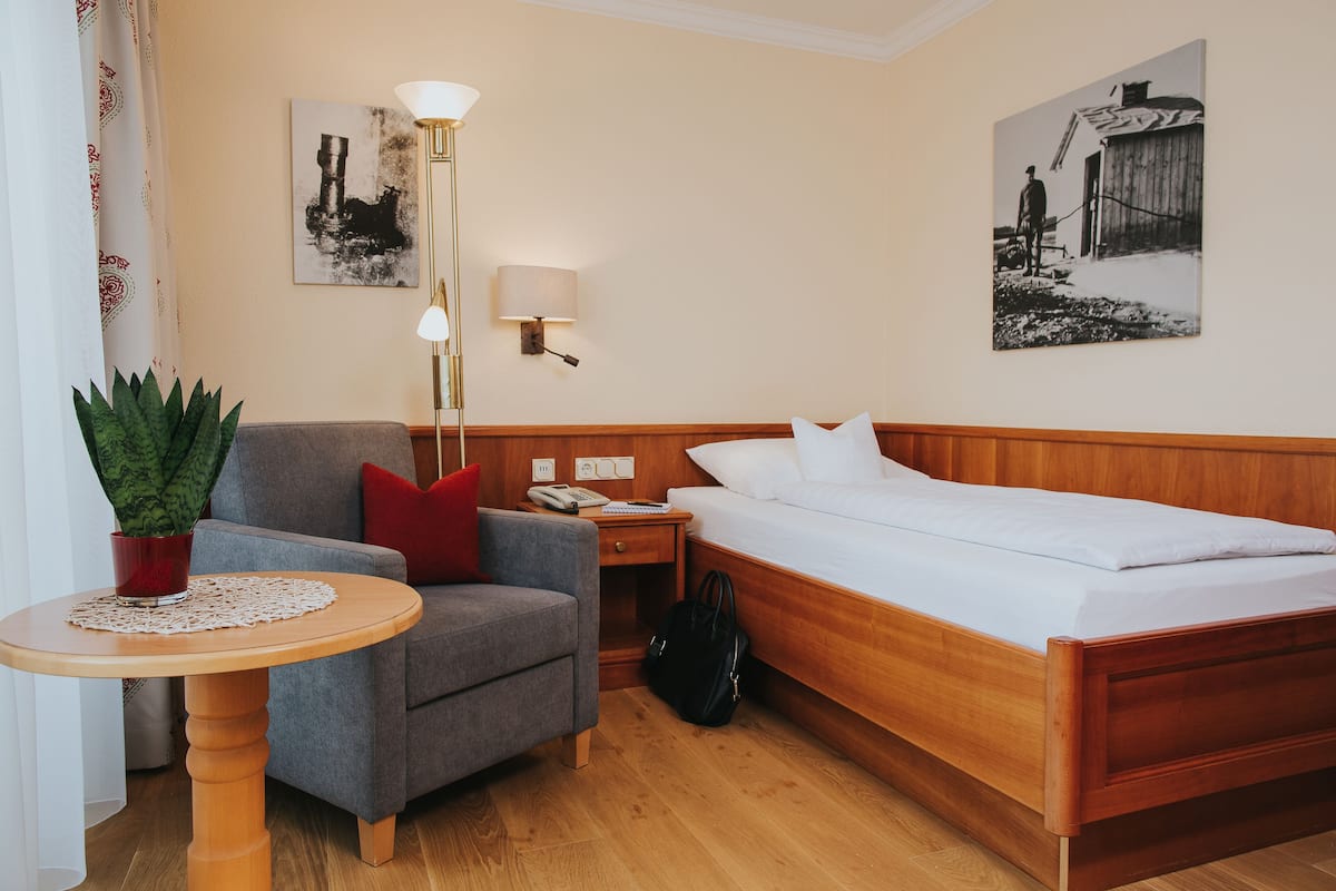 A hotel room with a wooden bed, a grey armchair, a small table with a plant, a lamp, and a wall-mounted phone.