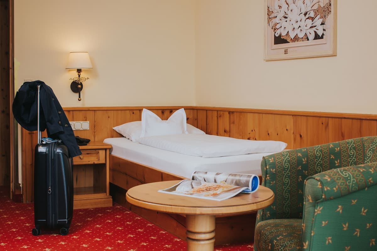 A hotel room with a bed, a wooden nightstand, a lamp, a green patterned armchair, a round wooden table, and a black suitcase.