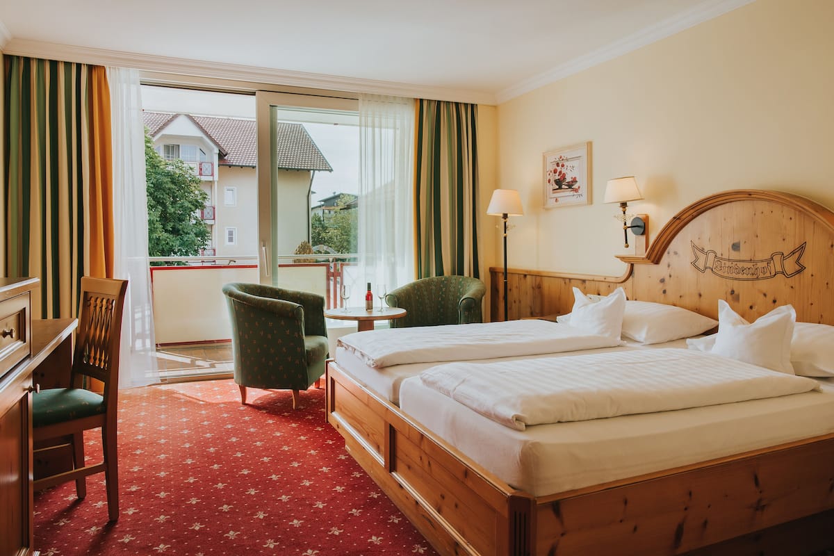 A hotel room with two beds, a desk, a chair, a small table with a bottle, and a view of a building through a window.