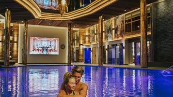 An indoor swimming pool with a modern design, featuring a large screen displaying a sports event in the background.