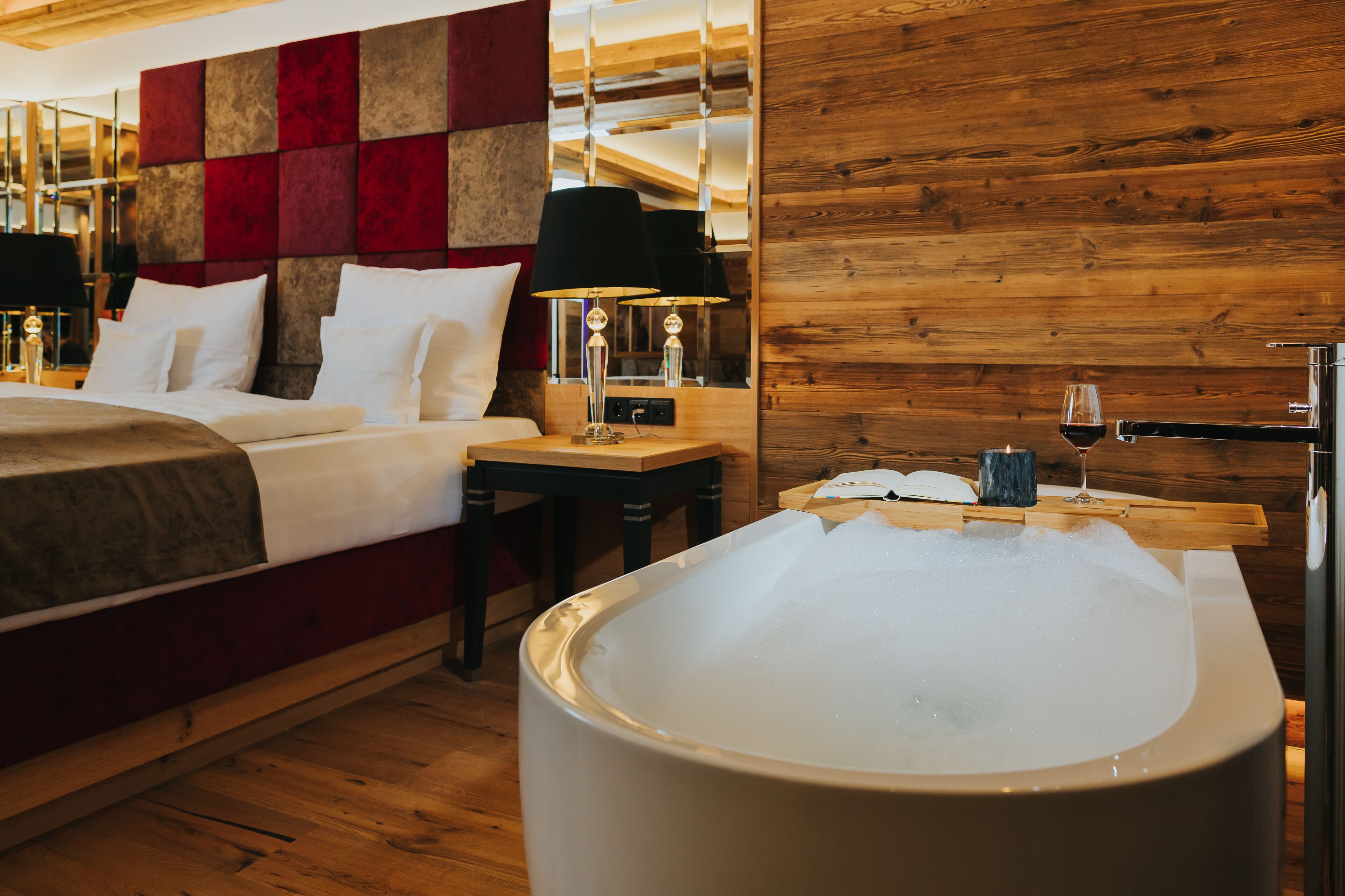 A modern hotel room with a large bed, a wooden headboard, a wooden wall, a small table with a lamp, and a hot tub.