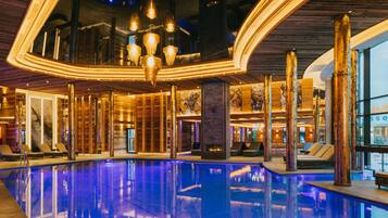 Un área de piscina interior lujosa con un diseño moderno, que cuenta con una gran piscina, zonas de estar y una estructura de techo singular.