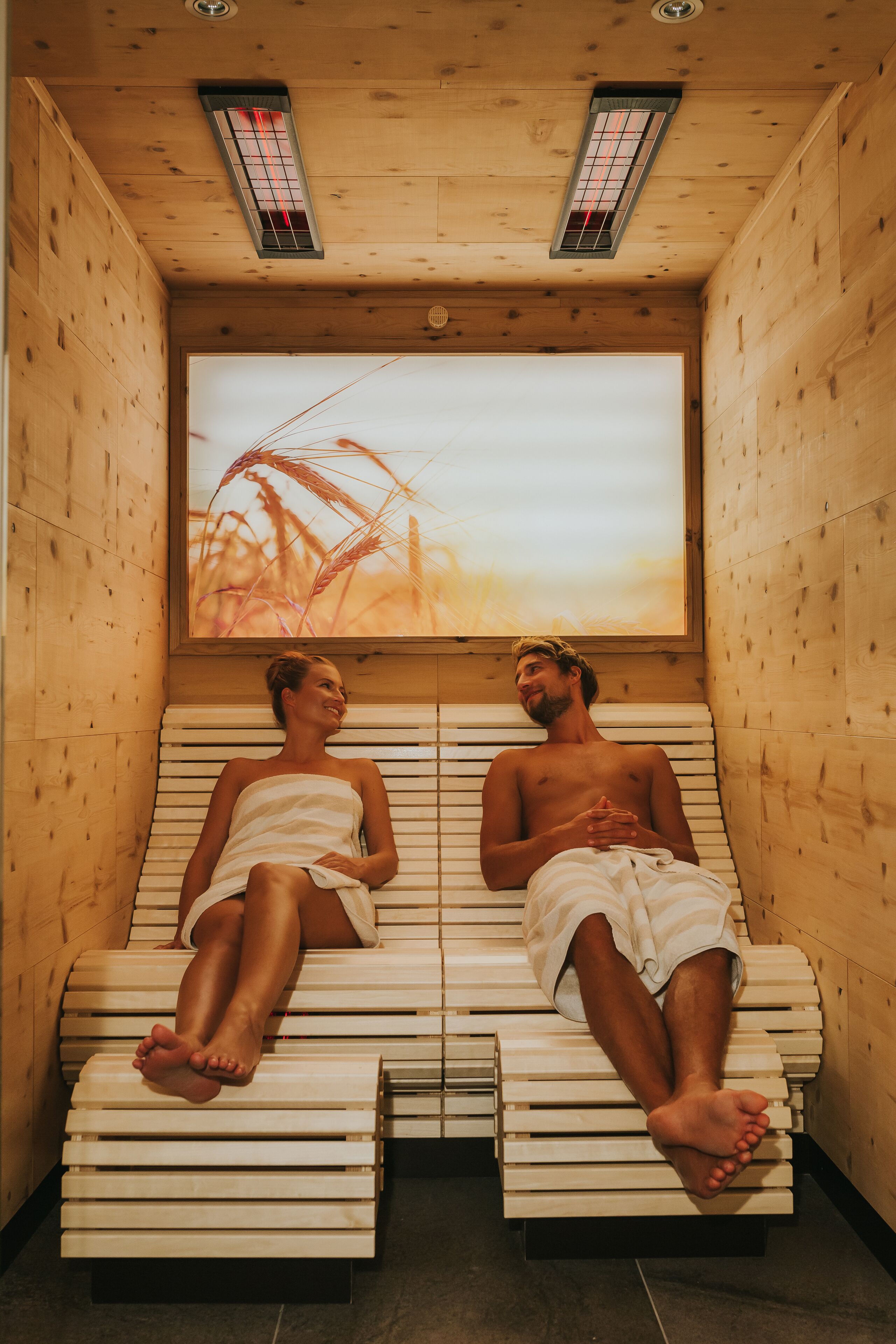 Dos personas en una sauna con paredes y bancos de madera, una ventana con vista a un campo de trigo, y una lámpara en el techo.