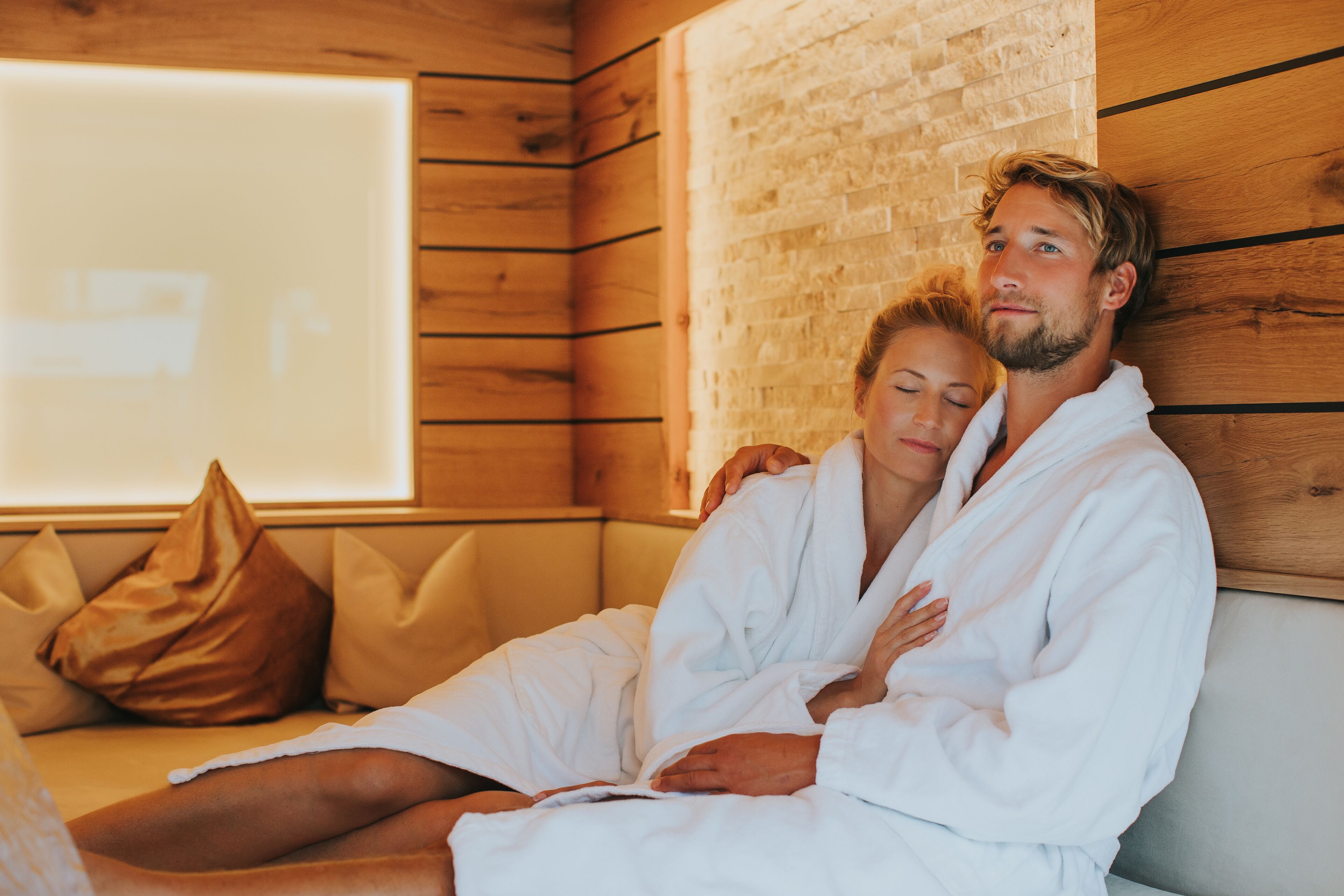 A couple in white robes sitting on a sofa in a room with wooden walls and a large window.
