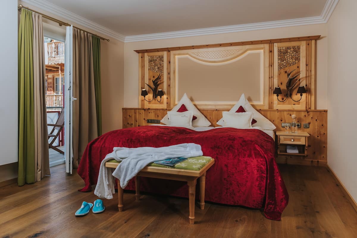 A bedroom with a wooden headboard, a red bedspread, a wooden bench with a towel, and a pair of shoes on the floor.
