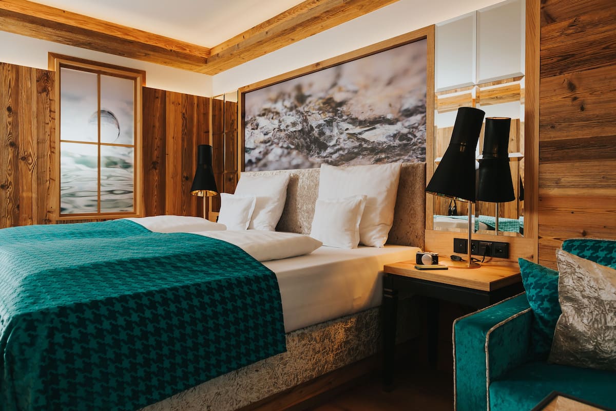 A bedroom with a large bed, a wooden headboard, a nightstand with a lamp, and a view of the outdoors through a window.