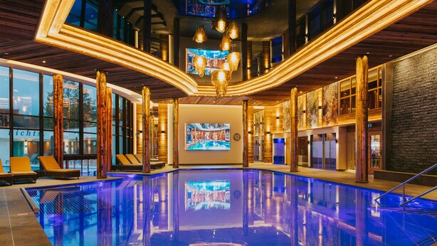 A modern indoor swimming pool with a curved design, large windows, and a central digital display. - Ortners Resort (Bad Fuessing)