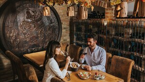 A wooden dining table with two people seated, surrounded by rustic chairs. A large wooden barrel with intricate carvings is visible in the background. - Ortners Resort (Bad Fuessing)