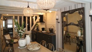 Een eetgedeelte met een houten tafel, stoelen en een vaas met bloemen. Er is een keuken met donkere kasten en een spoelbak. Een grote spiegel hangt aan de muur, en een kroonluchter aan het plafond.