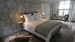 Een slaapkamer met een groot bed, twee nachtkastjes met lampen, een stoel, een raam met gordijnen en een bont tapijt op de vloer.