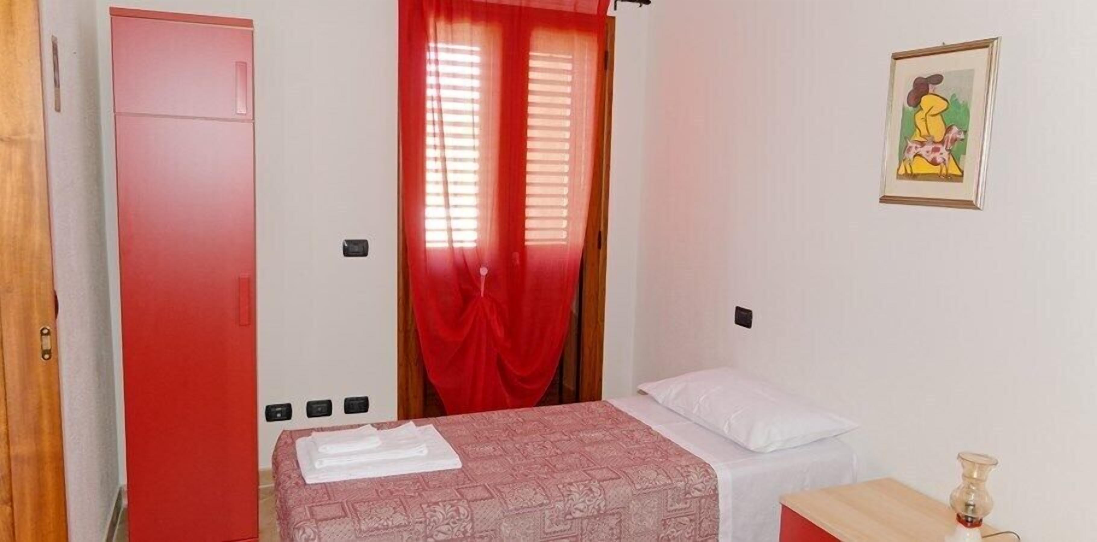 Un dormitorio con cama, una puerta roja, una ventana con cortinas rojas y un cuadro en la pared.