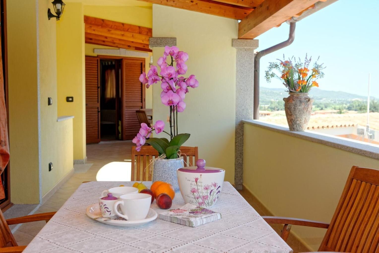 A balcony with a table set for breakfast, featuring a vase of pink flowers, a fruit bowl, and a coffee cup.