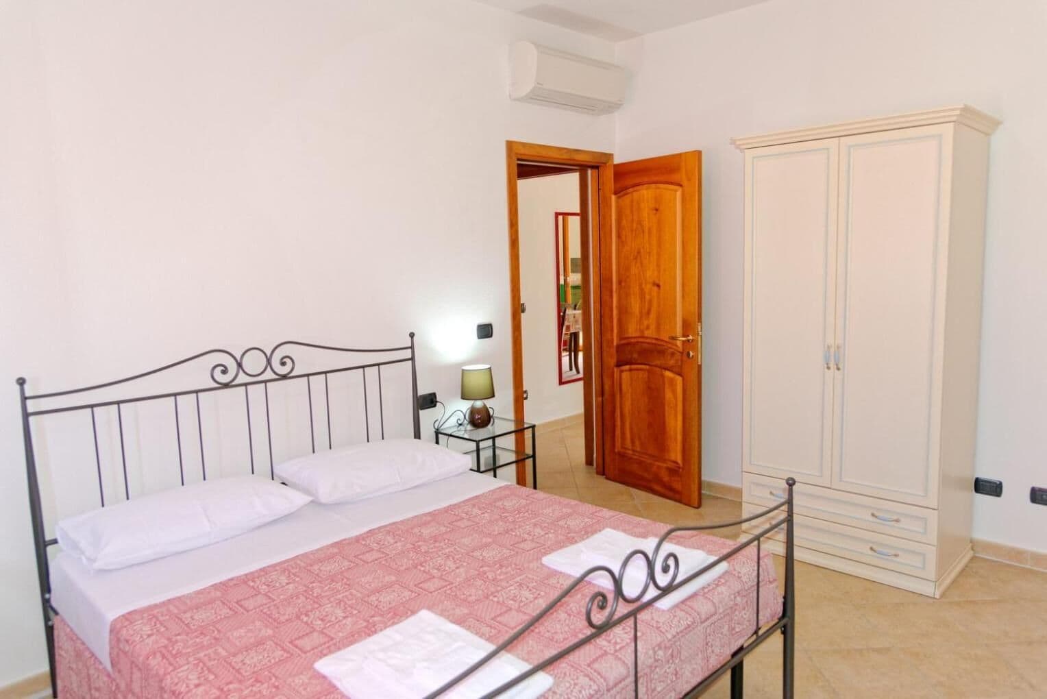 A bedroom with a bed, a nightstand, a lamp, a wardrobe, and a door leading to another room.