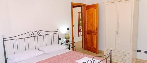 A bedroom with a bed, a nightstand, a lamp, a wardrobe, and a door leading to another room.