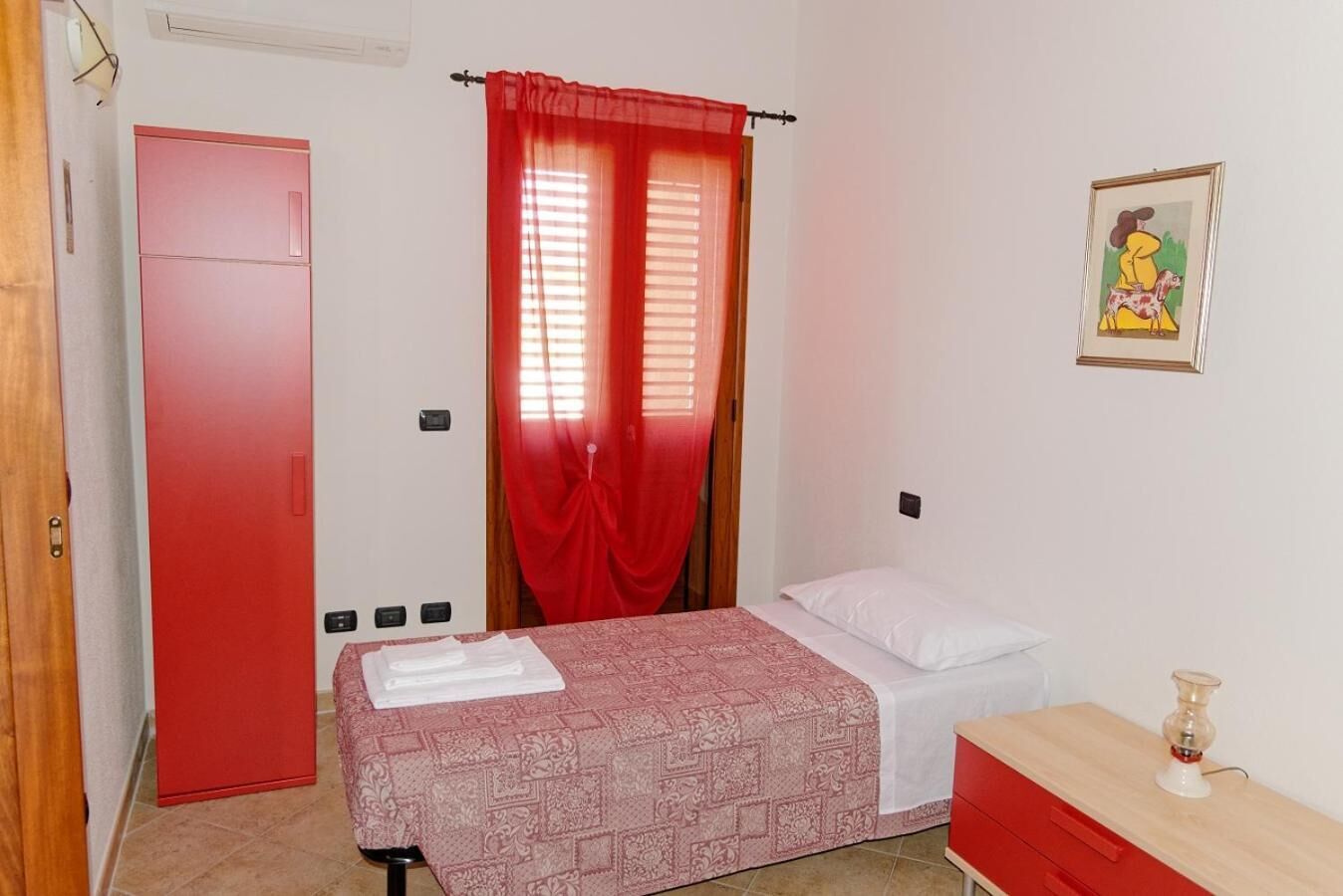 A bedroom with a bed, a nightstand, a red wardrobe, a red door, and a window with red curtains.