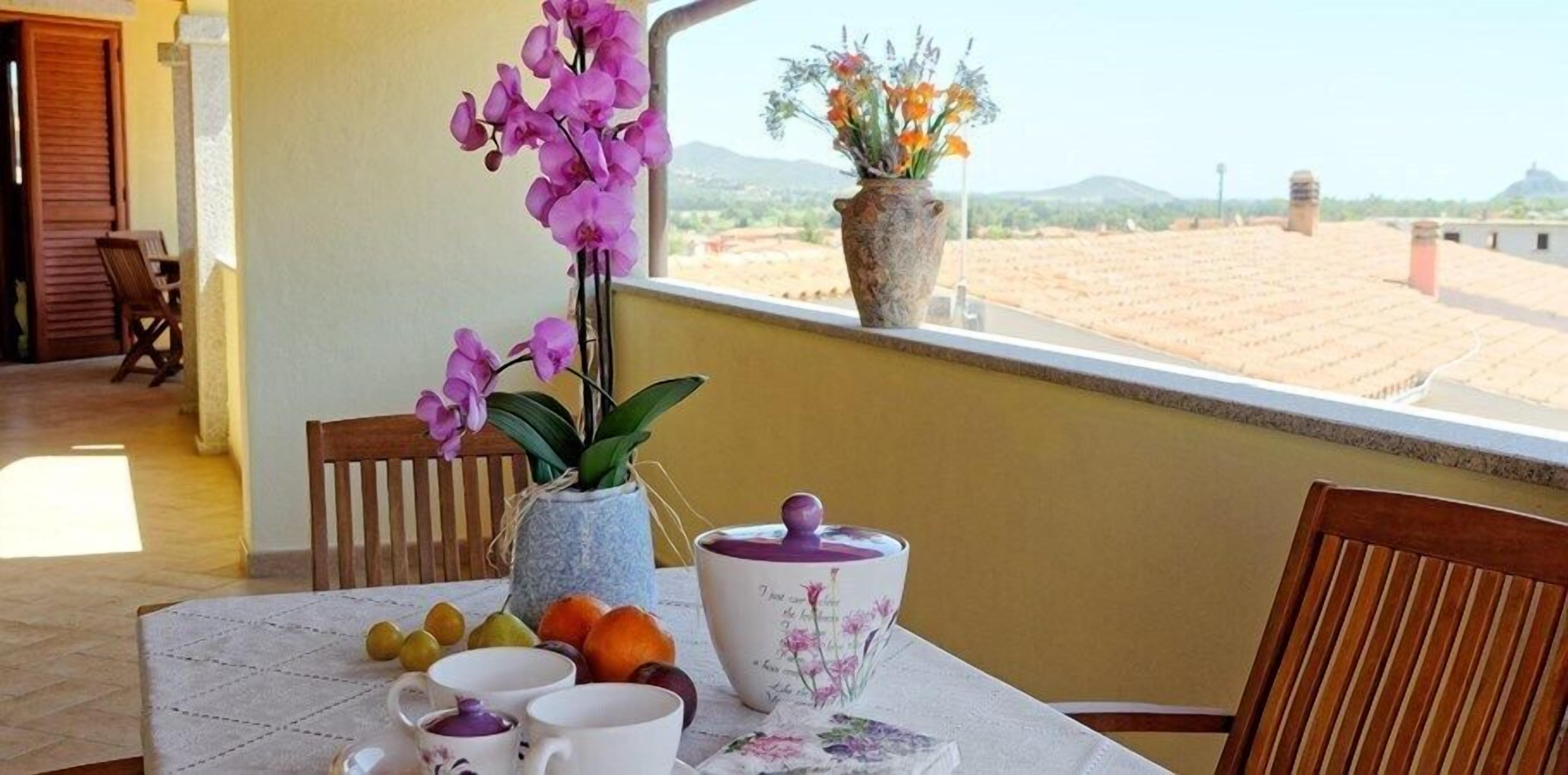 Ein Balkon mit Tisch, gedeckt für Tee, mit einer Vase mit lila Blumen, einer Obstschale und einer dekorativen Teekanne.