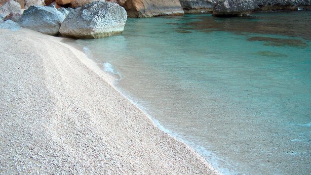Een strand met helder turquoise water, grote rotsen en een rotsachtige klif op de achtergrond.
