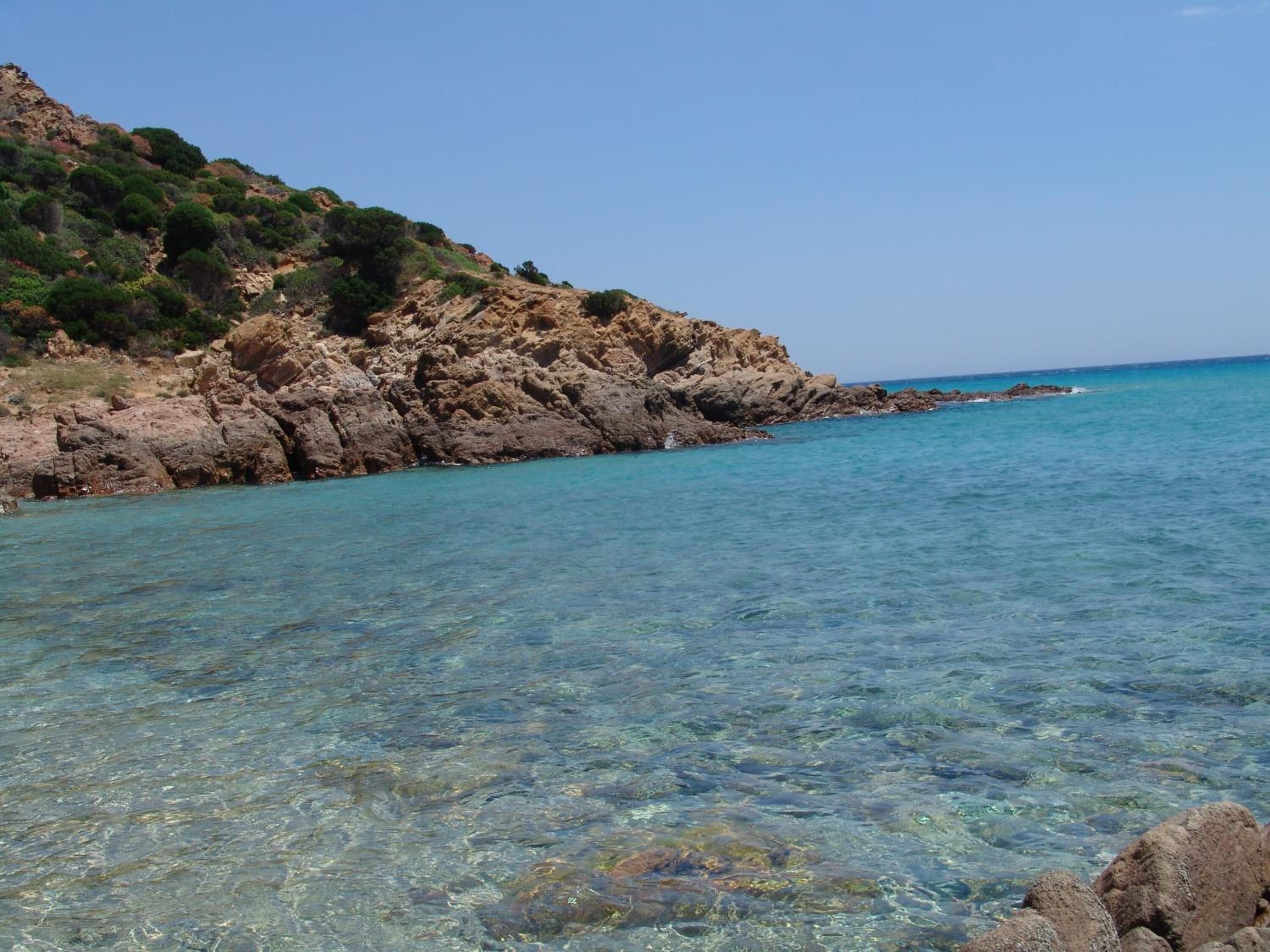 A coastal landscape with clear turquoise water, rocky cliffs, and sparse vegetation.