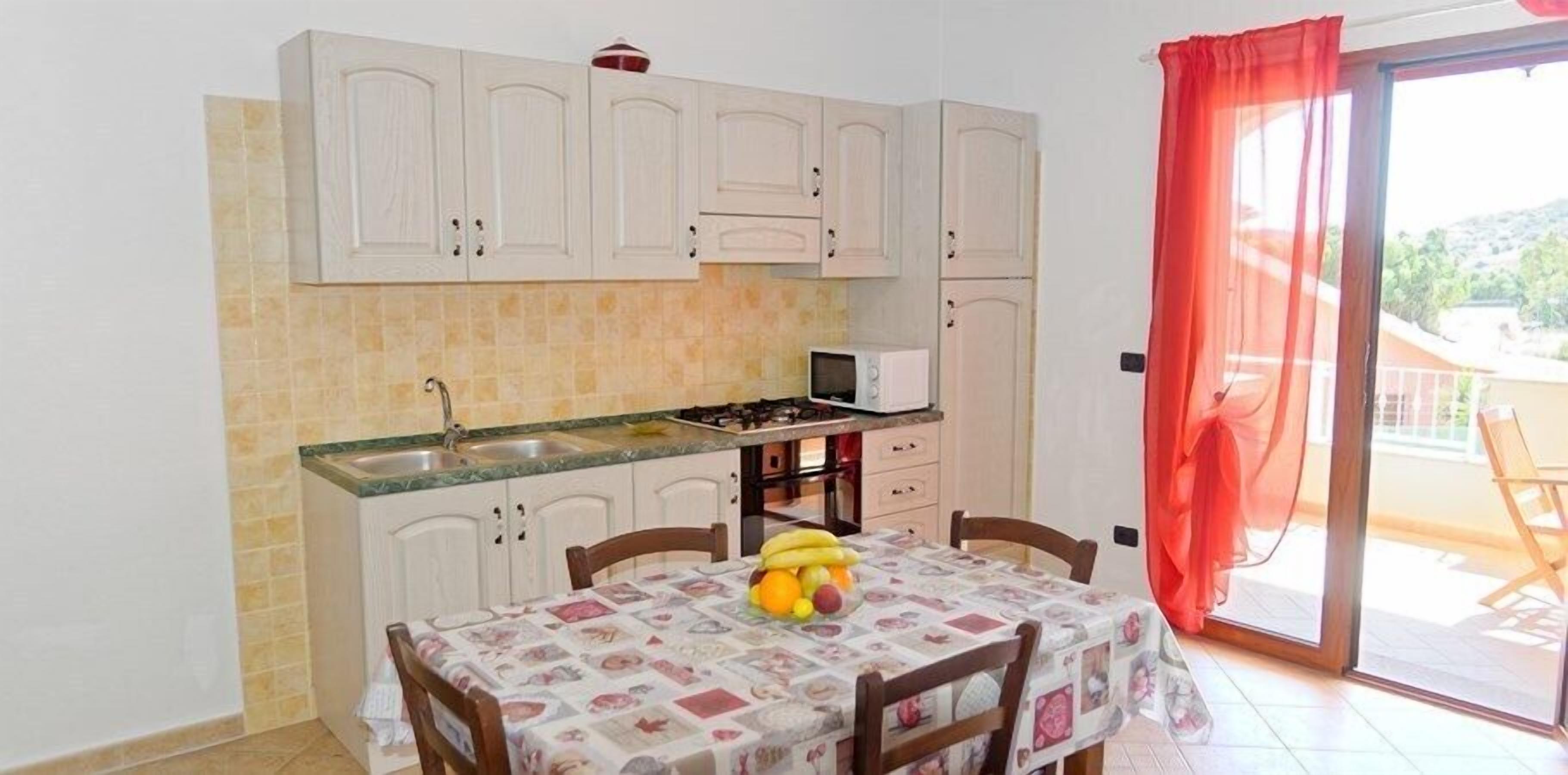 Una cocina compacta con armarios blancos, un microondas y una estufa a gas. Hay una mesa de comedor con mantel y fruta encima.