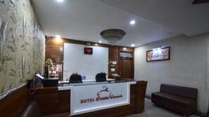 A hotel reception area with a reception desk, a digital clock, a chandelier, a framed picture on the wall, and a sofa.