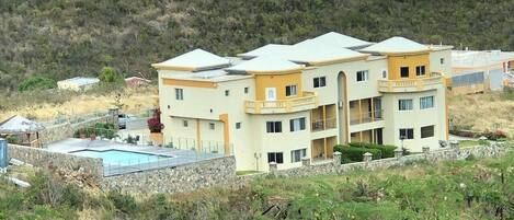 A multi-story building with a swimming pool, surrounded by greenery and hills.
