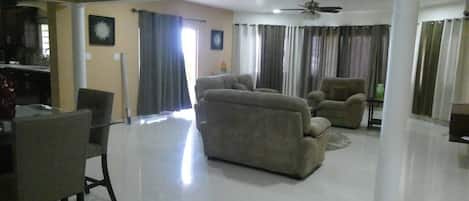 A spacious living room with a sofa set, a dining table with chairs, and a ceiling fan.