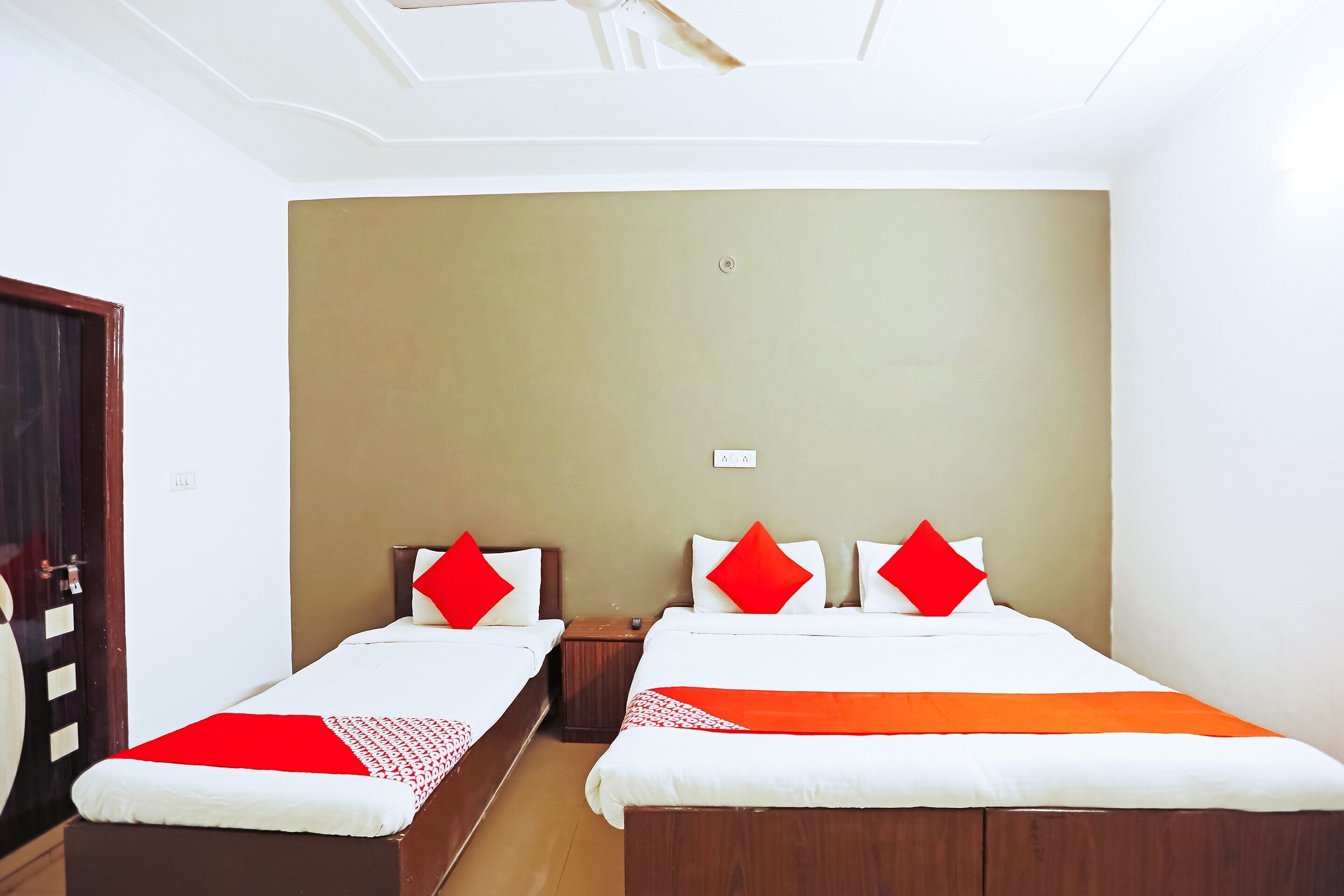 A hotel room with two single beds, each with a red pillow and a white bedspread. There is a wooden nightstand between the beds, and the room has beige walls and a white ceiling.