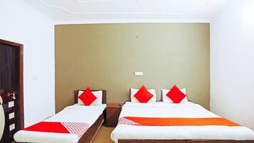 A hotel room with two single beds, each with a red pillow and a white bedspread. There is a wooden nightstand between the beds, and the room has beige walls and a white ceiling.