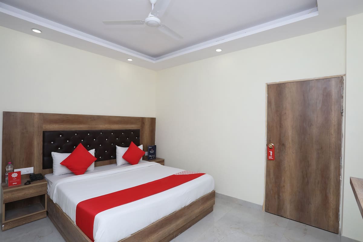 Une chambre d’hôtel avec un grand lit, une tête de lit en bois, une couvre-lit rouge et blanche, une table de chevet avec un téléphone et un objet rouge, et une porte en bois avec une pancarte rouge.