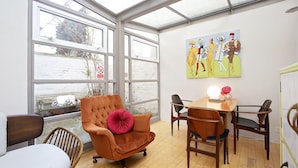 Una habitación con un ventanal grande, una mesa de comedor con sillas, un sillón con reposeras y una obra de arte colgada en la pared.
