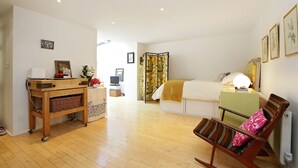 Una habitación con piso de madera, una cama con manta amarilla, un sillón mecedor con almohada rosa, una mesita de noche de madera con una lámpara, y un estante en la pared con una cesta.