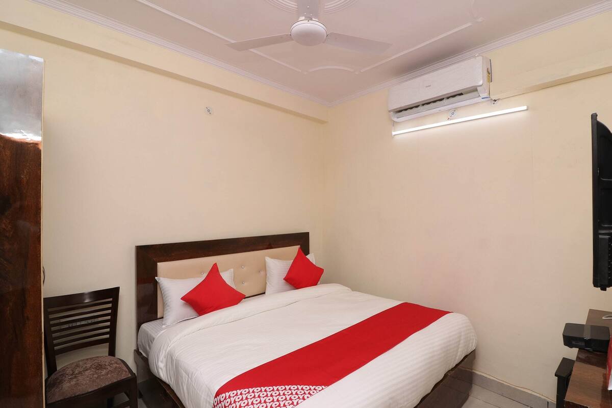 Habitación de hotel con una cama grande, cabecera de madera, colcha roja y blanca y un equipo de aire acondicionado en la pared.
