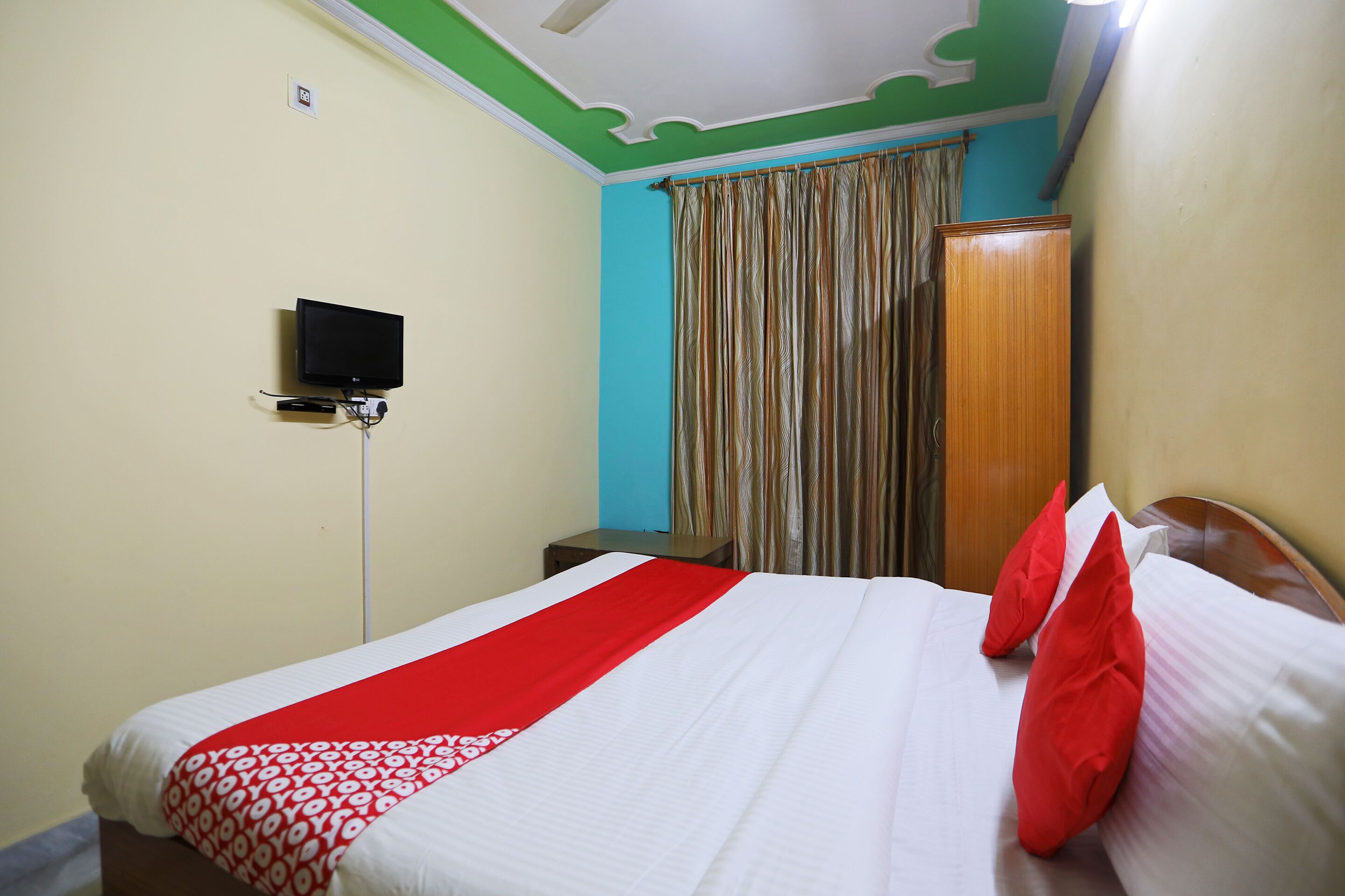 A neatly made bed with a red and white bedspread, a television on a stand, and a wooden wardrobe in a room with teal walls and a green ceiling.