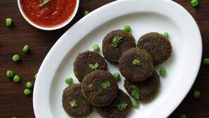 Un plato blanco con varias hamburguesas redondas y oscuras, adornadas con guisantes verdes y perejil, acompañado de un pequeño bol de salsa roja.