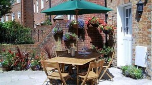 Eine Terrasse mit Holztisch und -stühlen, einem grünen Sonnenschirm und hängenden Blumenkörben.