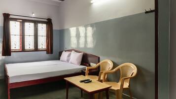 A single-bed room with a wooden table, two plastic chairs, a ceiling fan, and a window with curtains.