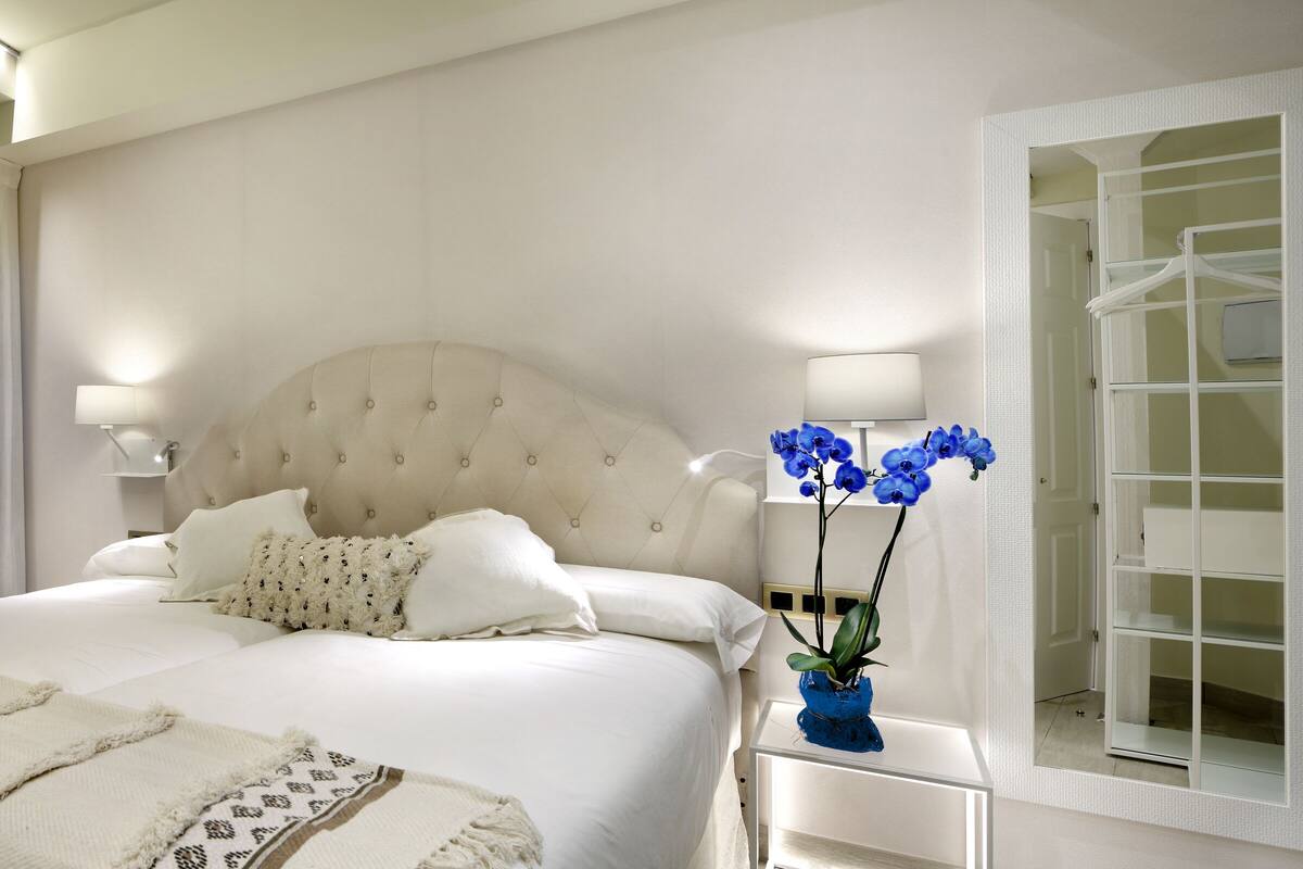 A bedroom with a large bed, white bedding, a tufted headboard, a bedside table with a lamp, and a blue orchid vase.