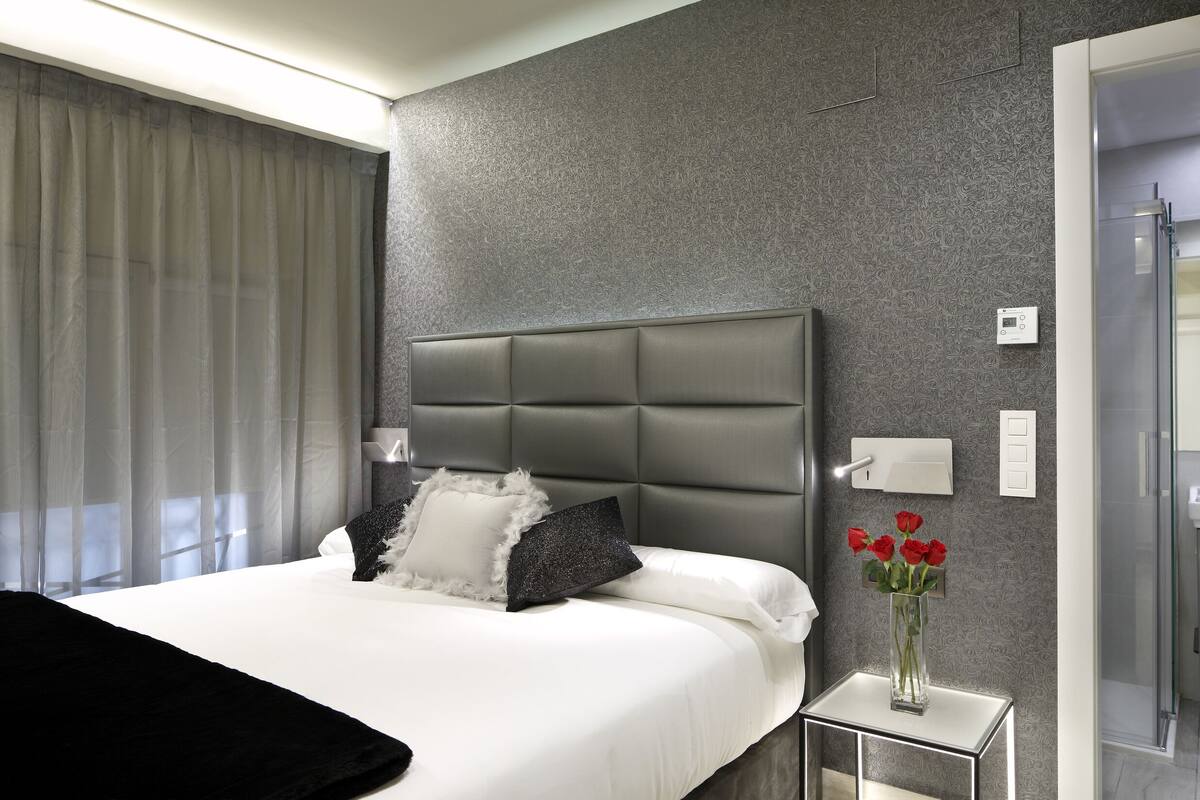 A modern hotel room with a large bed, a bedside table with a vase of flowers, and a bathroom visible through an open door.