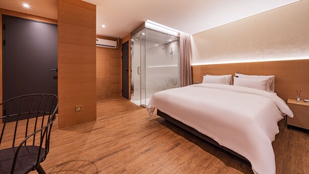 Una habitación de hotel moderna con una cama grande, una zona de estar con una silla, un vestidor y un box de ducha de vidrio.
