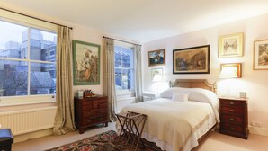 Une chambre à coucher avec un grand lit, une commode, une table de chevet, une fenêtre avec des rideaux et plusieurs tableaux encadrés accrochés au mur.