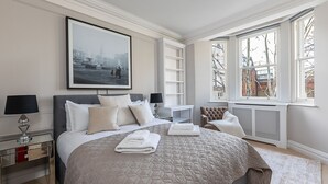 A bedroom with a large bed, a nightstand, a reading chair, a window with a view of buildings, and a radiator. - JOIVY Astonishing flats near Mayfair & Piccadilly Circus (London)