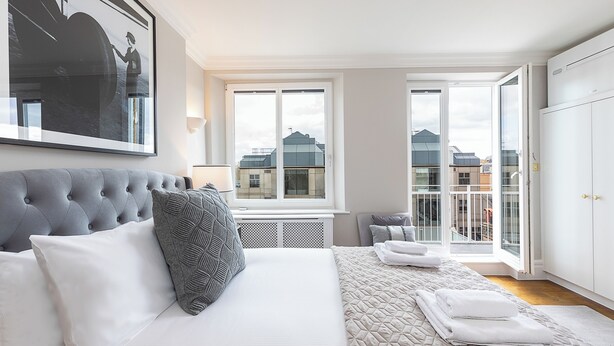 A bedroom with a large bed, a grey tufted headboard, a window with a view of buildings, and a white wardrobe. - JOIVY Astonishing flats near Mayfair & Piccadilly Circus (London)