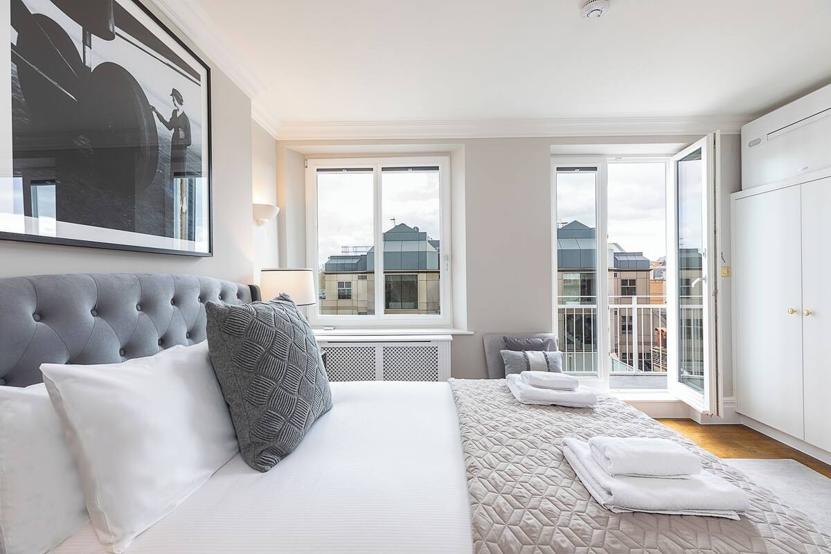 Un dormitorio con una cama grande, un cabecero tapizado de color gris, una ventana con vistas a edificios y un armario blanco.